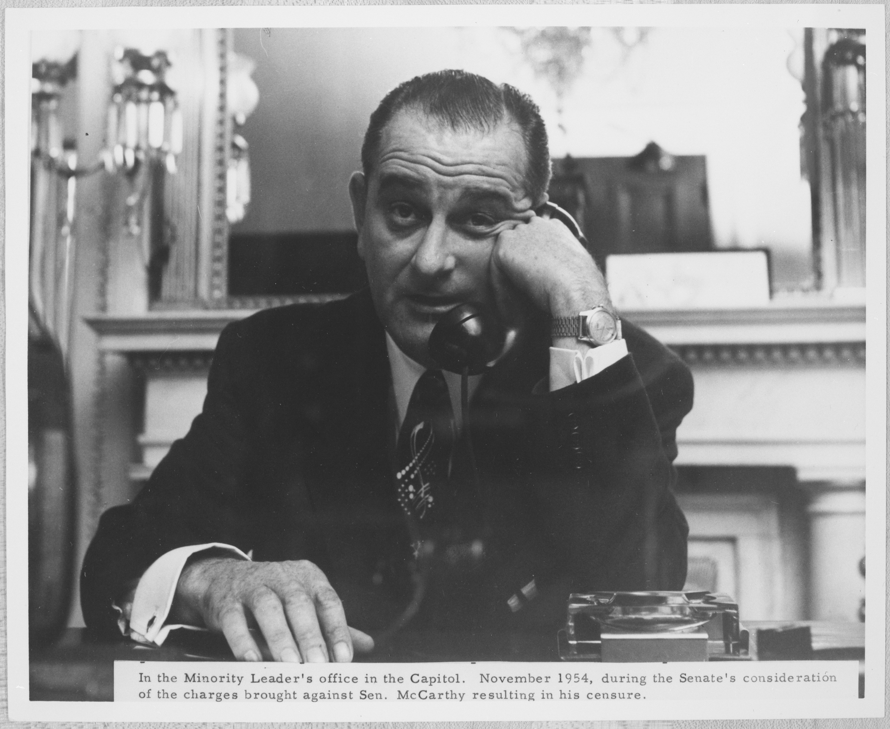 Lyndon Baines Johnson raises his eyebrows as he talks on the phone, seated behind a desk. 